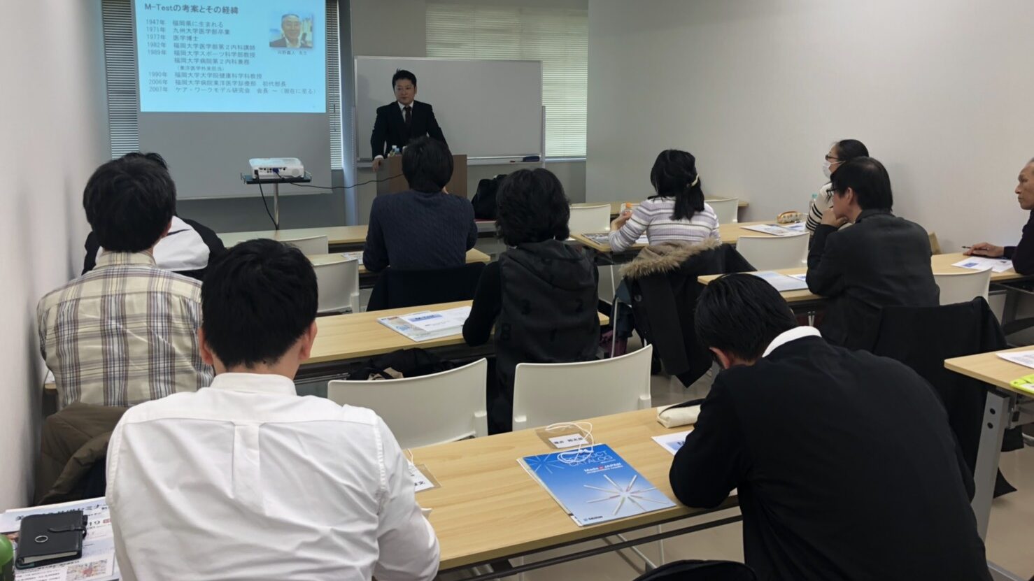 有名な技術について解説する講師と、それを真剣に聴講する参加者たちの様子。おるきの整体が行う代替医療の学術的な教育活動を表現したセミナー風景。